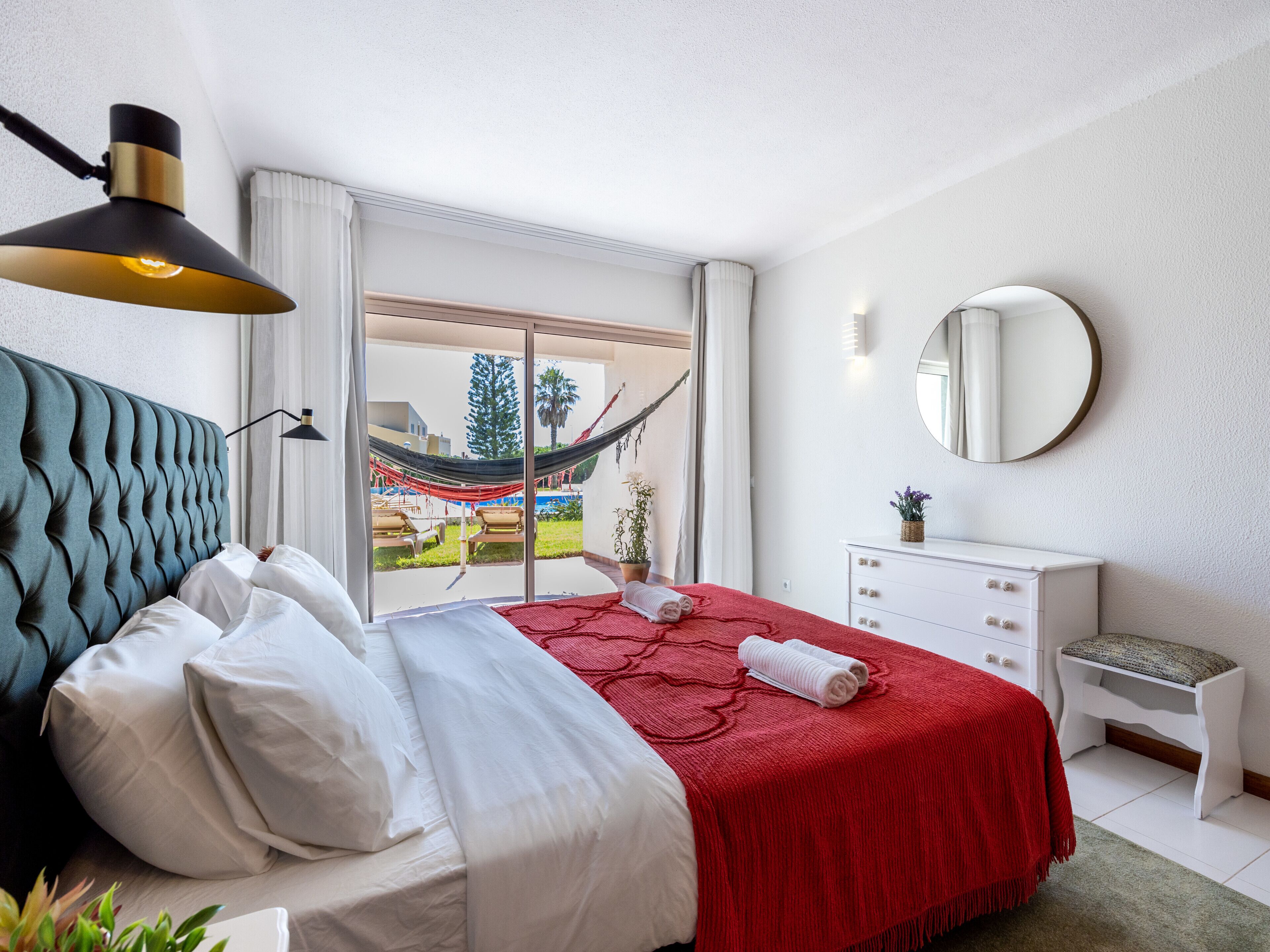 Ein Schlafzimmer mit Bett, Nachttischlampe, Kommode, Spiegel und Blick ins Freie.
