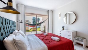 Ein Schlafzimmer mit Bett, Nachttischlampe, Kommode, Spiegel und Blick ins Freie.