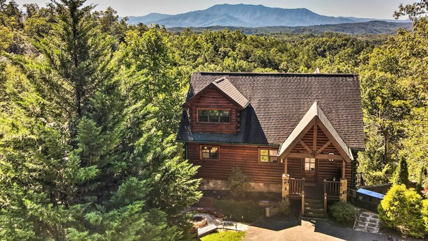 A log cabin with a steeply pitched roof, surrounded by dense forest and mountains in the background.