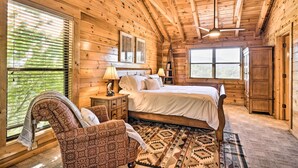 A wooden cabin bedroom with a bed, two bedside tables, a chair, and a large window with blinds.