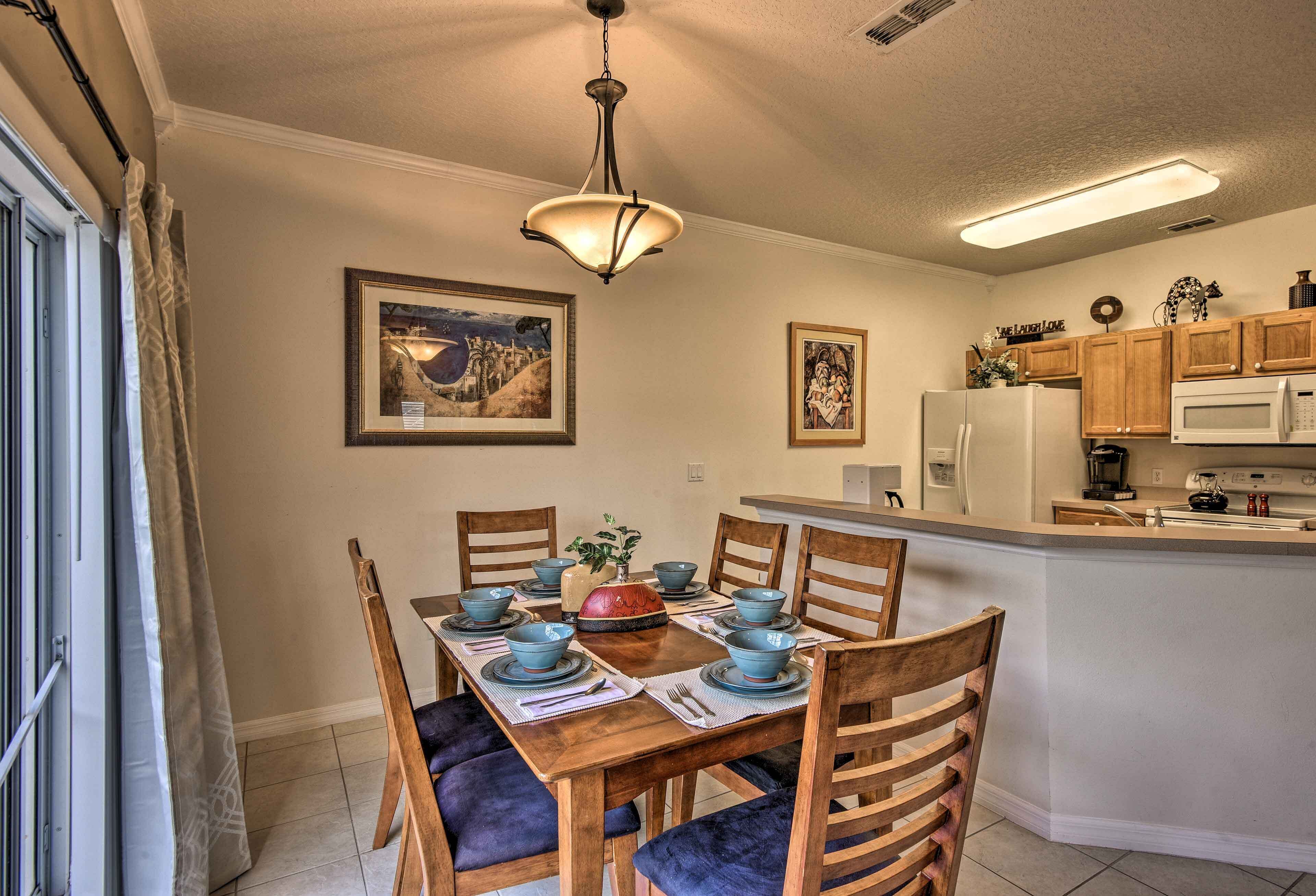 A dining area with a wooden table set for six, surrounded by chairs with cushioned seats. A chandelier hangs above the table, and artwork is displayed on the walls.