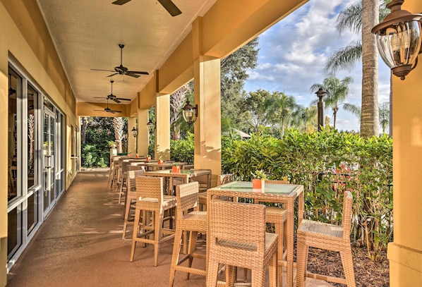 An outdoor patio with wicker tables and chairs, ceiling fans, and a view of palm trees and greenery.