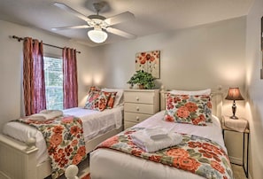 A bedroom with two beds, a ceiling fan, a window with curtains, a bedside lamp, and a floral painting on the wall.