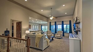 A spacious living room with a grey sofa, two armchairs, a coffee table, and a TV stand. A wooden staircase with a railing leads to another room. - Expansive Townhome w/ Game Room & Community Pool (Branson West)