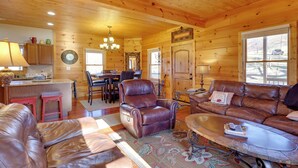 A cozy log cabin interior with a leather sofa, a leather armchair, a dining area with a chandelier, and a large window. - Decks, Panoramic Views! Mtn Retreat in Sparta (Sparta)