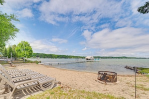 A beach with lounge chairs, a fire pit, and a dock extending into a lake.