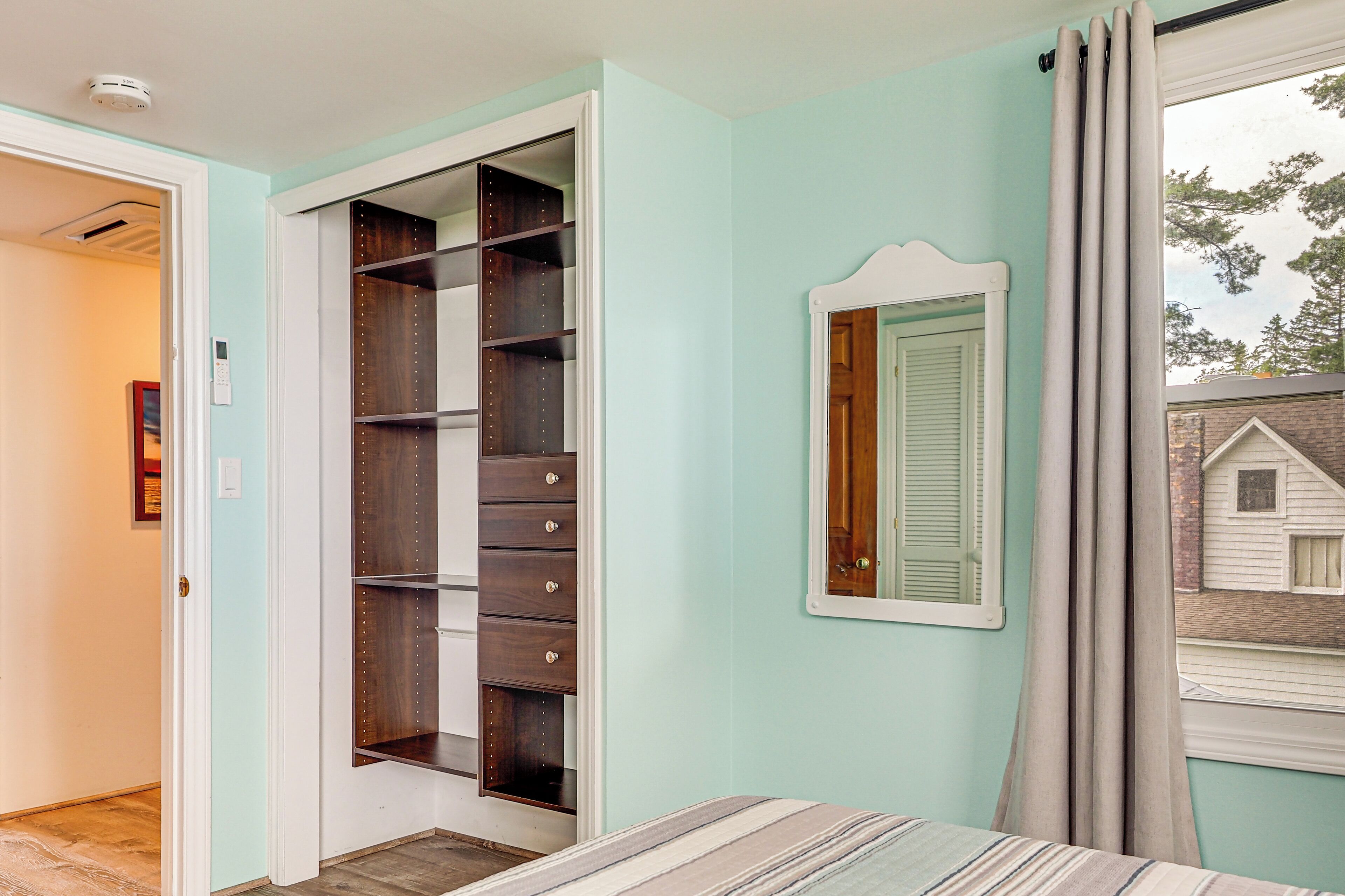 A bedroom with a bed, a walk-in closet, a window with curtains, and a view of a house outside.