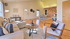 A spacious living room with a sectional sofa, a round coffee table, and a modern chair. A dining area with a brick accent wall and a kitchen in the background is also visible. - Pet-friendly Cottage With Game Room & Fire Pit! (Reva)