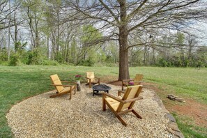 Un espace circulaire aménagé avec des chaises en bois et un foyer, entouré de gravier, avec un arbre au centre et un champ d’herbe en arrière-plan.
