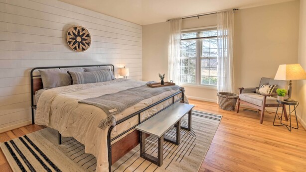 A bedroom with a bed, bedside table, lamp, bench, and a window with curtains. - Pet-friendly Cottage With Game Room & Fire Pit! (Reva)