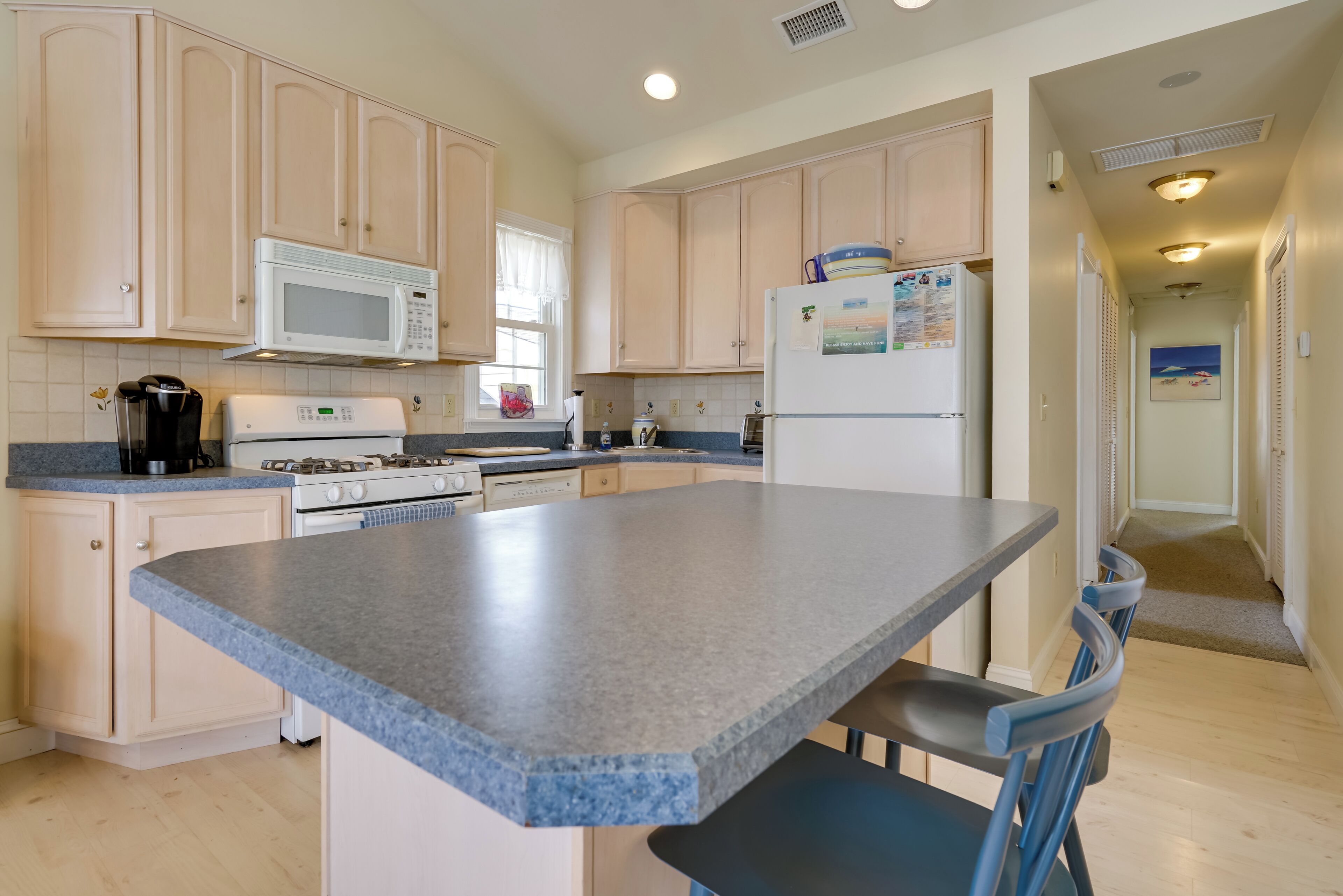 A kitchen with light wooden cabinets, a white microwave, a stove, and a refrigerator. There is a blue countertop and a chair.
