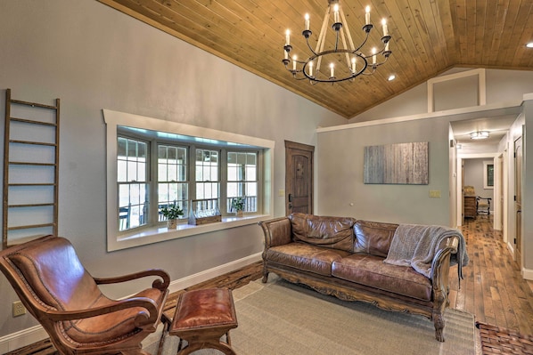 A spacious living room with a leather sofa, a wooden chair, a chandelier, and a large window with multiple panes.
