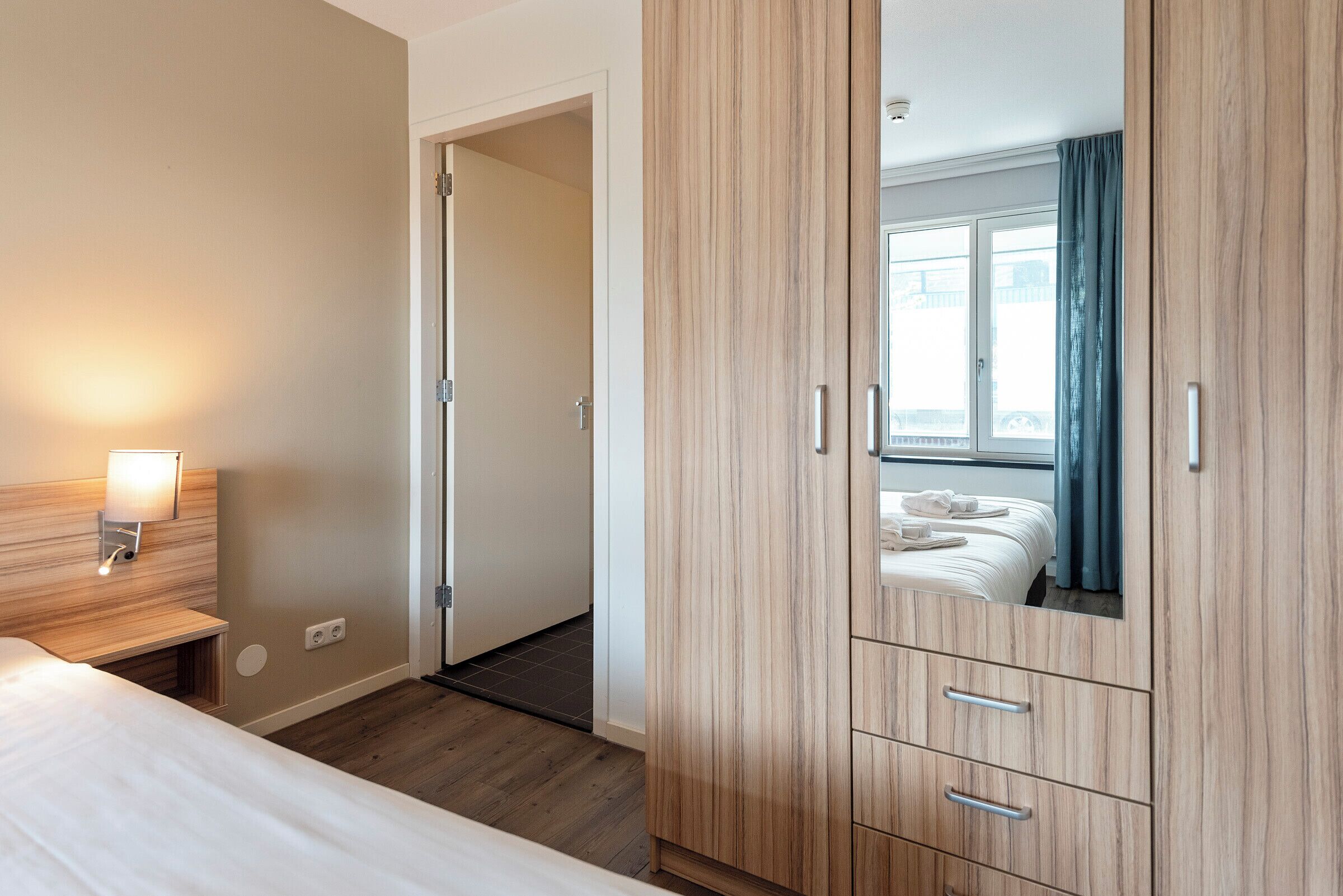 Een hotelkamer met een bed, een houten kledingkast, een nachtkastje met een lamp en uitzicht door een raam met gordijnen.
