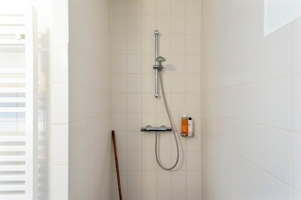 A shower with a handheld showerhead and a wall-mounted faucet, a wooden stick, and a small shelf with toiletries.