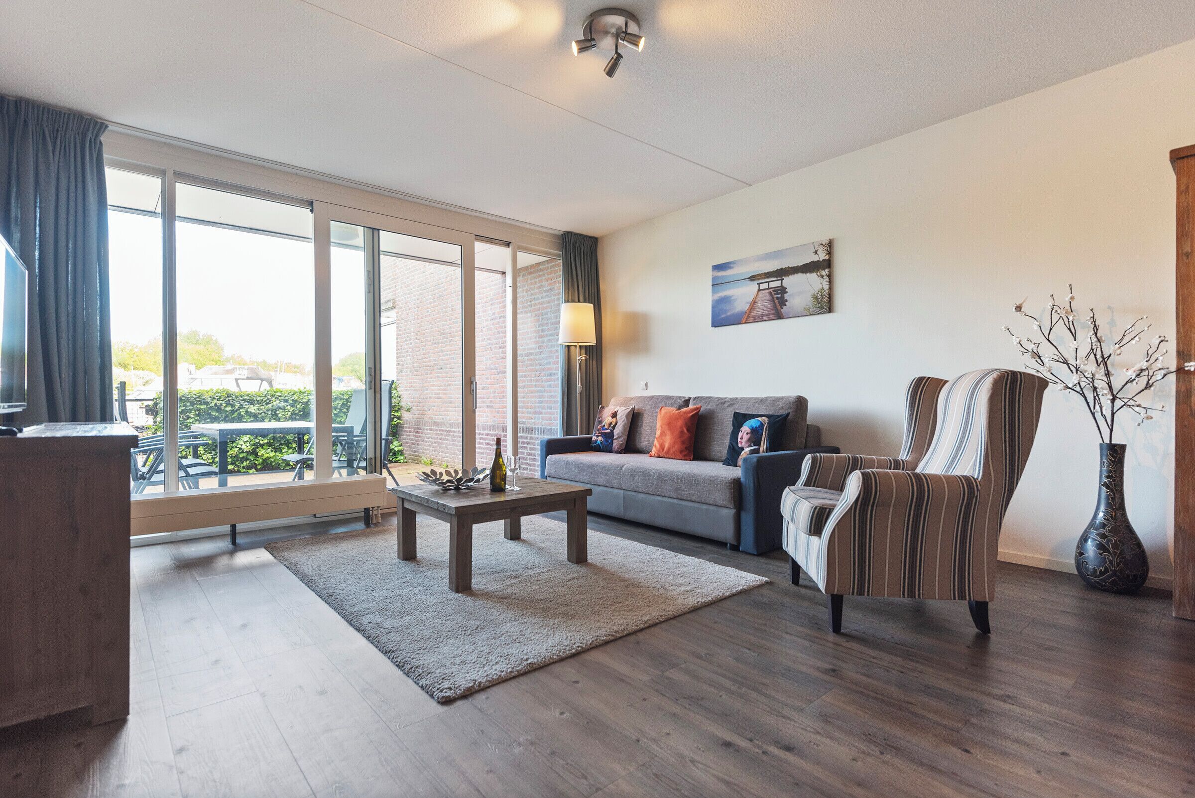 Een moderne woonkamer met een bank, fauteuil, salontafel en een groot raam met uitzicht op groen en een gebouw.