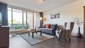 Een moderne woonkamer met een bank, fauteuil, salontafel en een groot raam met uitzicht op groen en een gebouw.