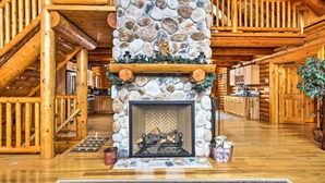 A log cabin interior with a stone fireplace, wooden staircase, and a kitchen area.