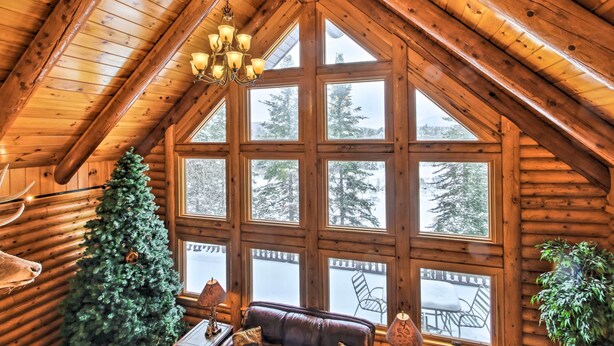 A log cabin interior with a large window, a Christmas tree, a leather sofa, and a chandelier.