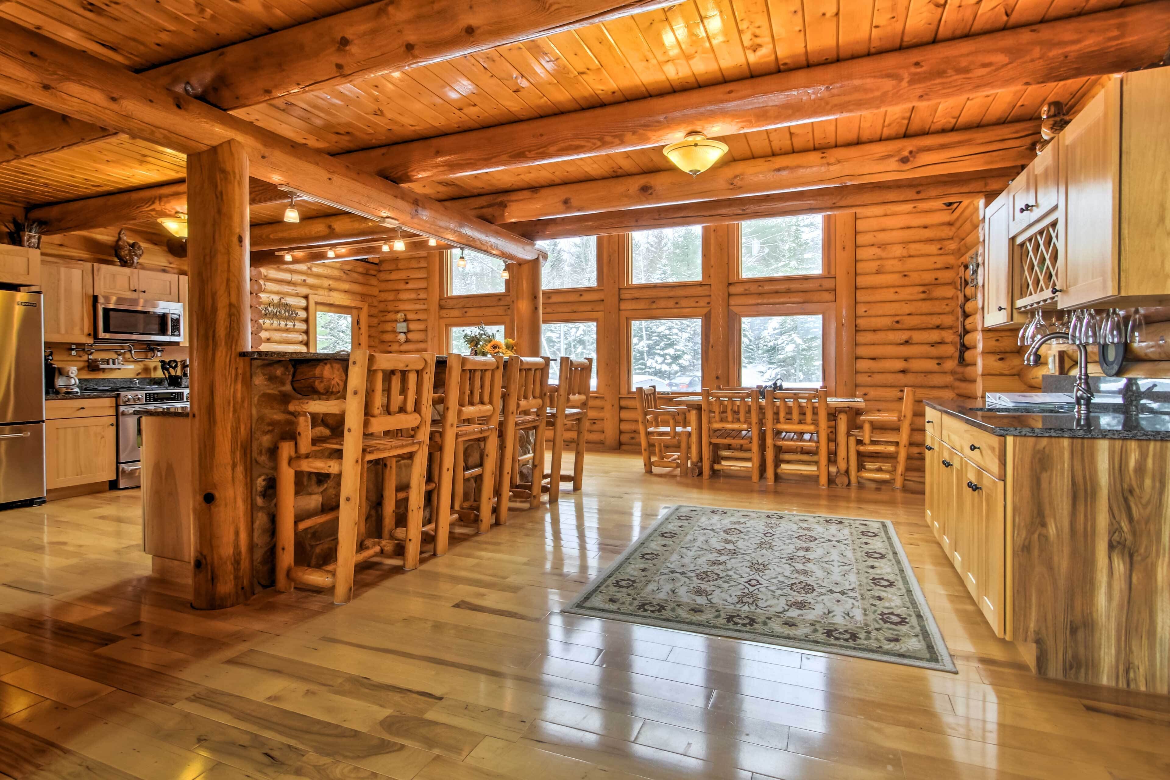 A log cabin kitchen with wooden cabinets, a dining area with chairs, a rug, and large windows.