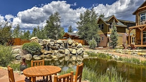 A wooden outdoor seating area with a pond and waterfall, surrounded by trees and a cabin-style building.