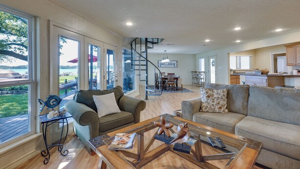 A spacious living room with a staircase leading to a dining area, large windows offering views of the outdoors, and a kitchen area. - Lakefront Montgomery Home w/ Porch & Dock! (Montgomery)