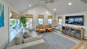 Ein geräumiges Wohnzimmer mit hoher Decke, einem großen Fernseher, einer Couch, zwei Sesseln, einer Holzkommode und einem Deckenventilator.