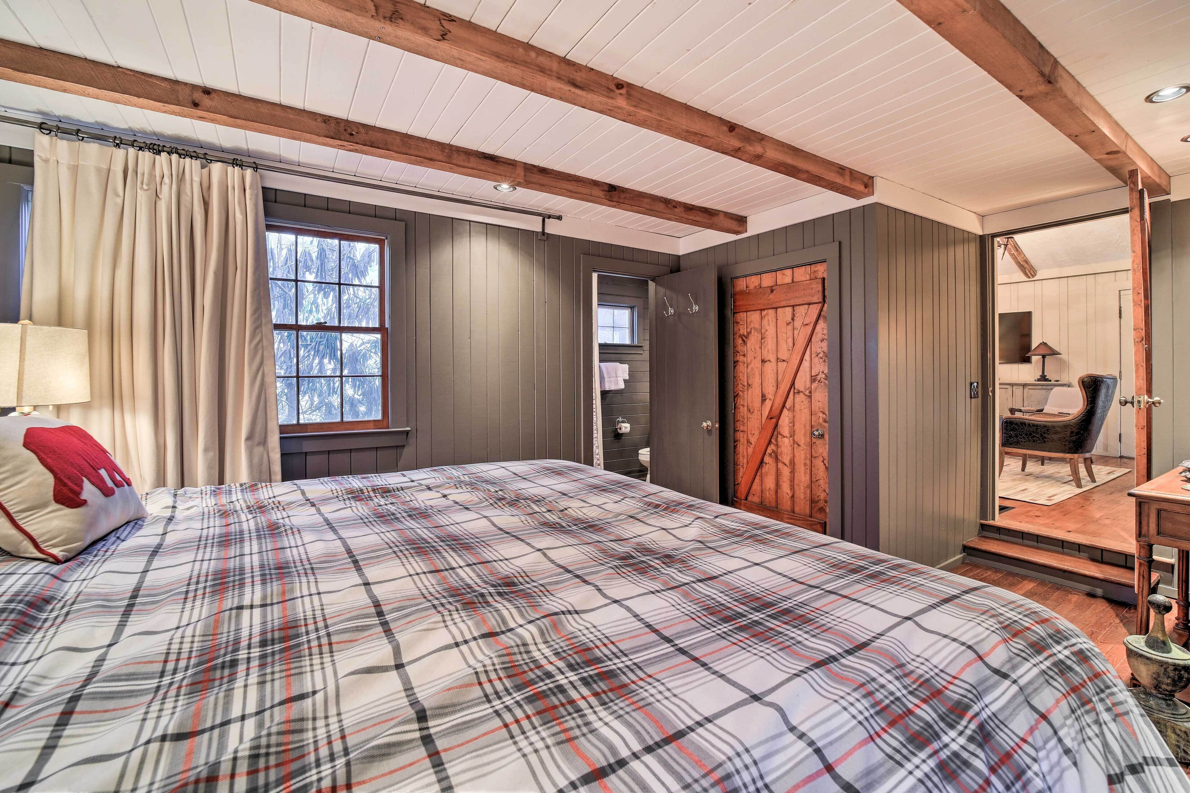 Un dormitorio con colcha a cuadros, vigas de madera, un ventanal grande y una puerta que da a un baño.