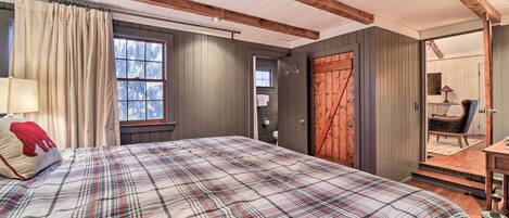 Un dormitorio con colcha a cuadros, vigas de madera, un ventanal grande y una puerta que da a un baño.