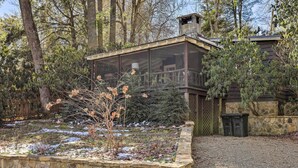 Un espace extérieur entouré d’arbres et d’un mur de pierre, avec une terrasse en bois et une structure fermée par des parois de verre.