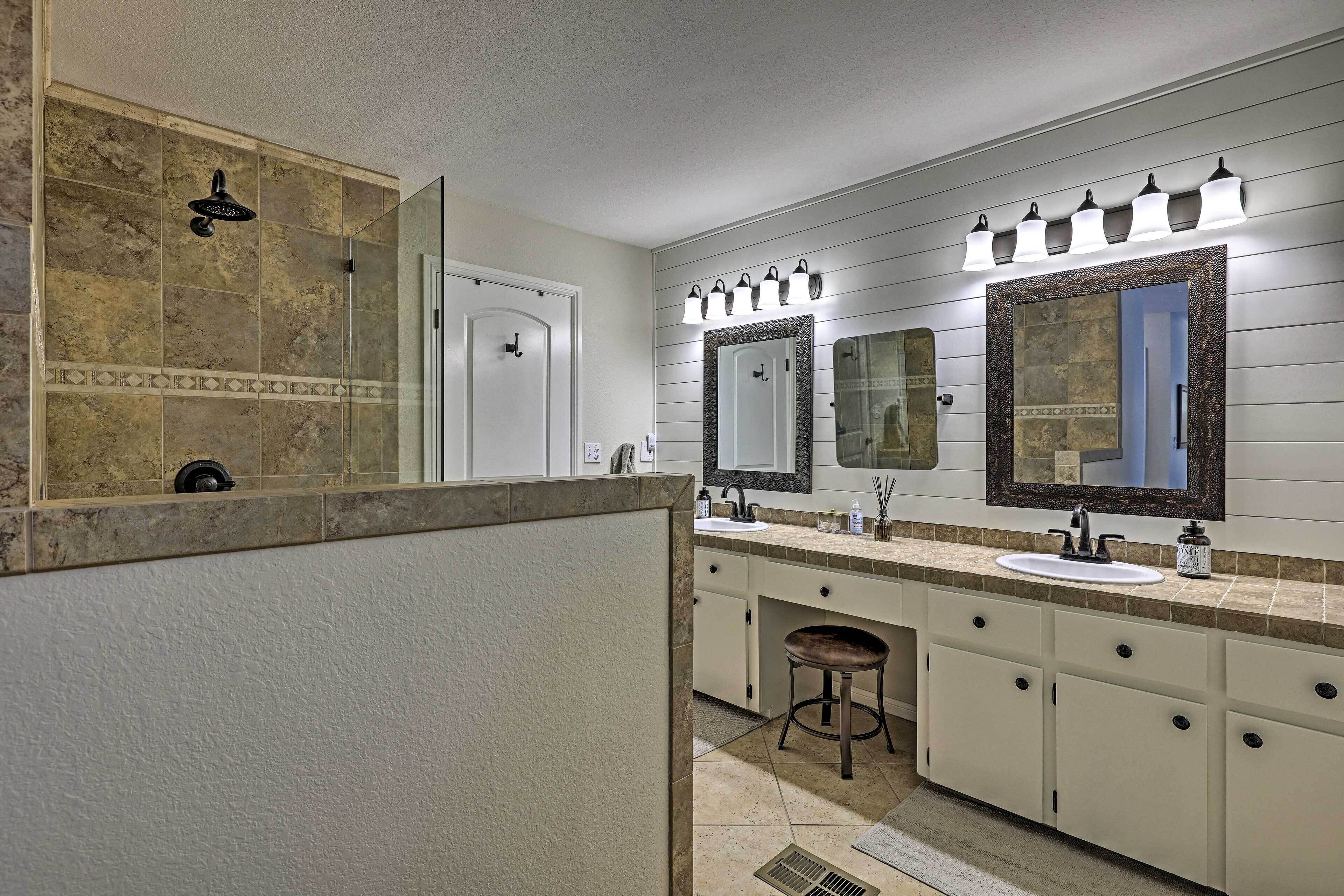 A bathroom with a large mirror, two sinks, a shower, and a vanity with a stool.
