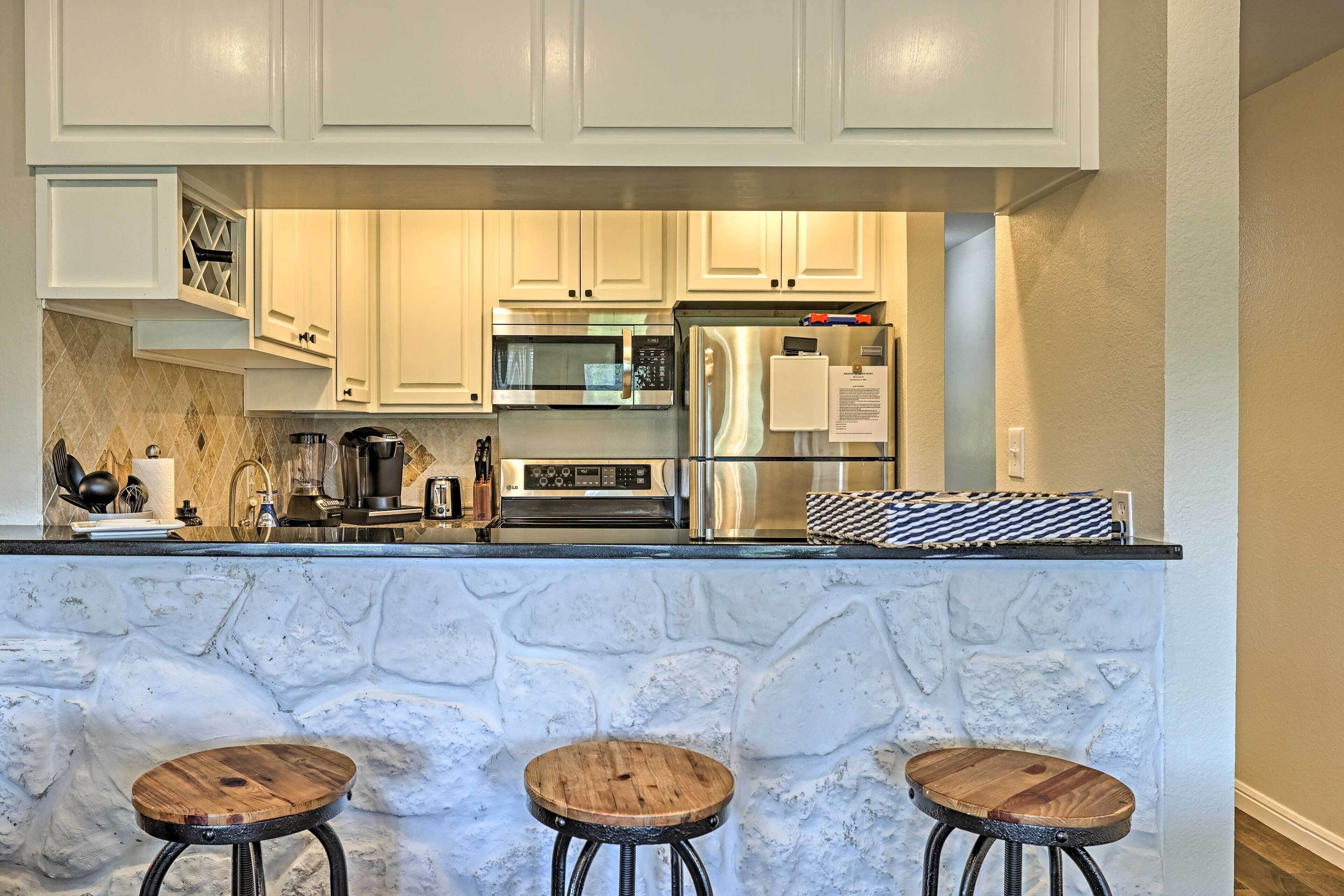 A kitchen with white cabinets, stainless steel appliances, a stone backsplash, and three wooden stools.