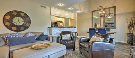 A modern living room with a sofa, armchairs, and a coffee table. There is a kitchen in the background with white cabinets and a dining area with a table and chairs.