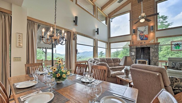 Un coin repas avec une table en bois dressée pour six personnes, entourée de sièges confortables. Une cheminée en pierre et de grandes fenêtres offrent une vue sur l’extérieur.
