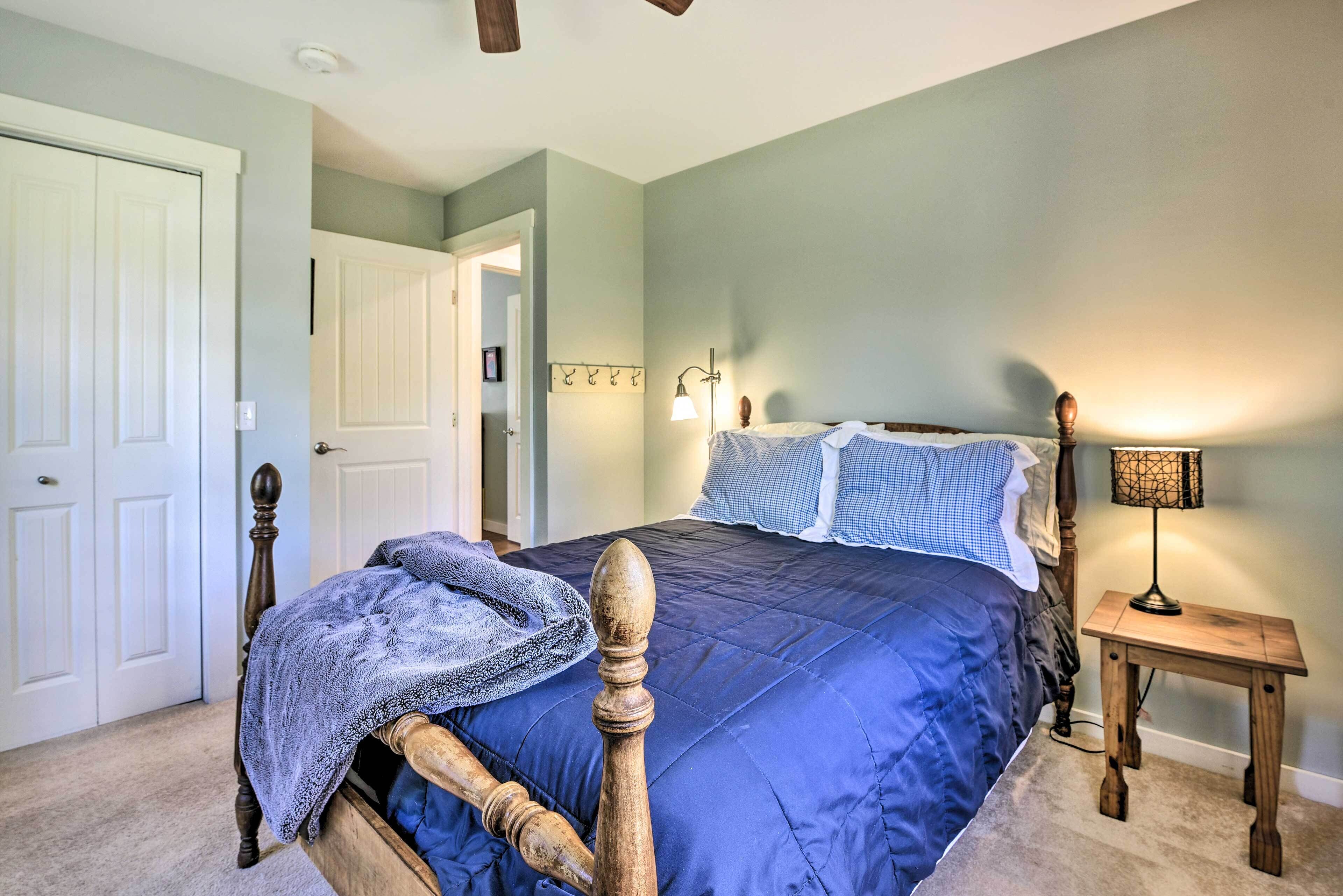 A bedroom with a wooden bed, a blue bedspread, a nightstand with a lamp, and a door leading to another room.