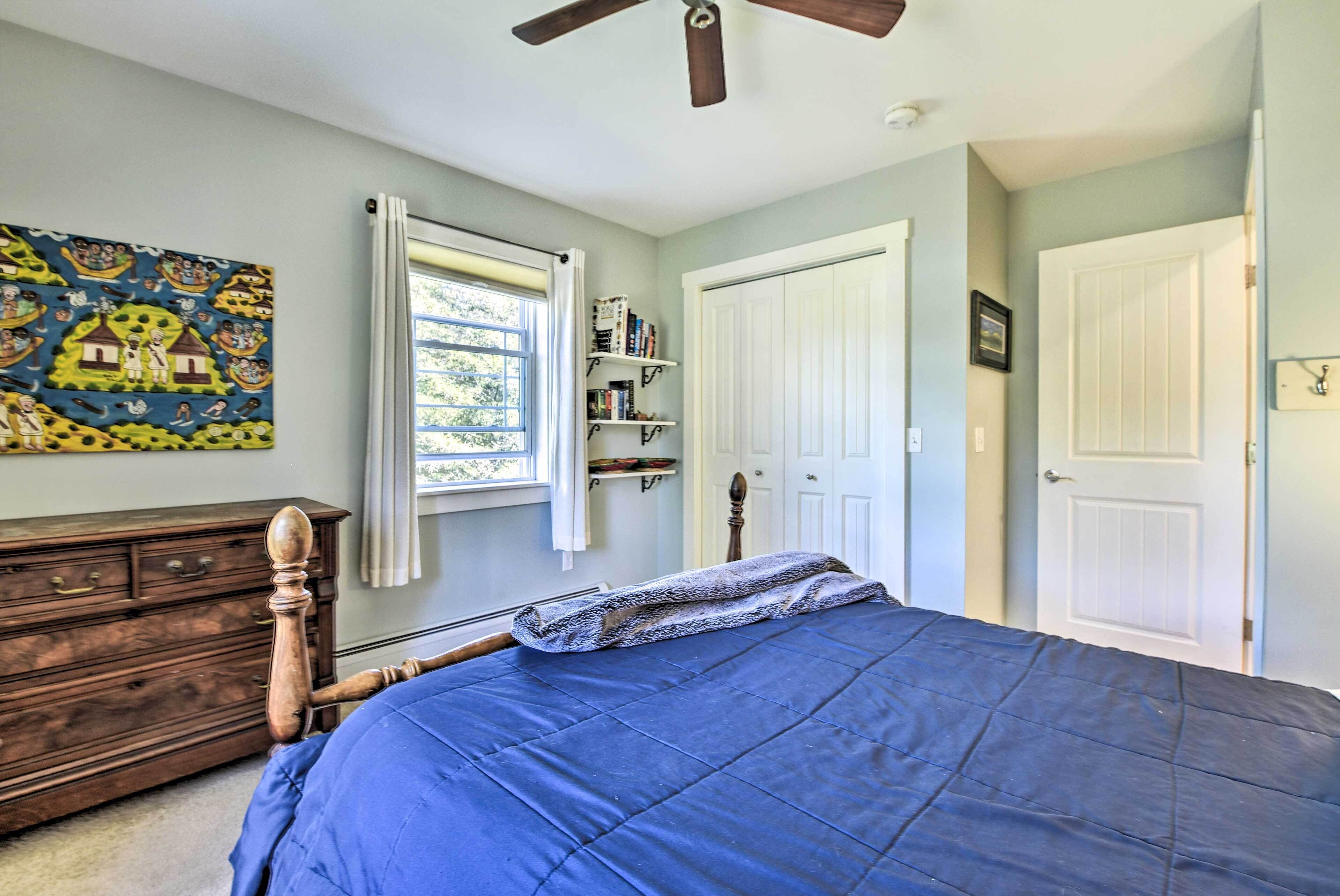 Un dormitorio con cama, cómoda de madera, ventana con cortinas, ventilador de techo y puerta.