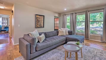 A living room with a grey sectional sofa, a small round table with a plant, and a large window with curtains.