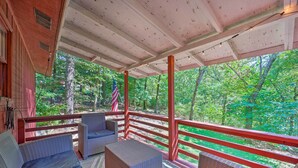 A covered porch with wicker furniture and a red railing, overlooking a forested area.