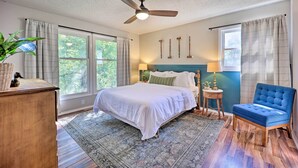 A bedroom with a bed, a blue armchair, a wooden dresser, a ceiling fan, and a window with curtains.