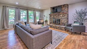 A living room with a stone fireplace, a gray sofa, and a view of the outdoors through large windows.