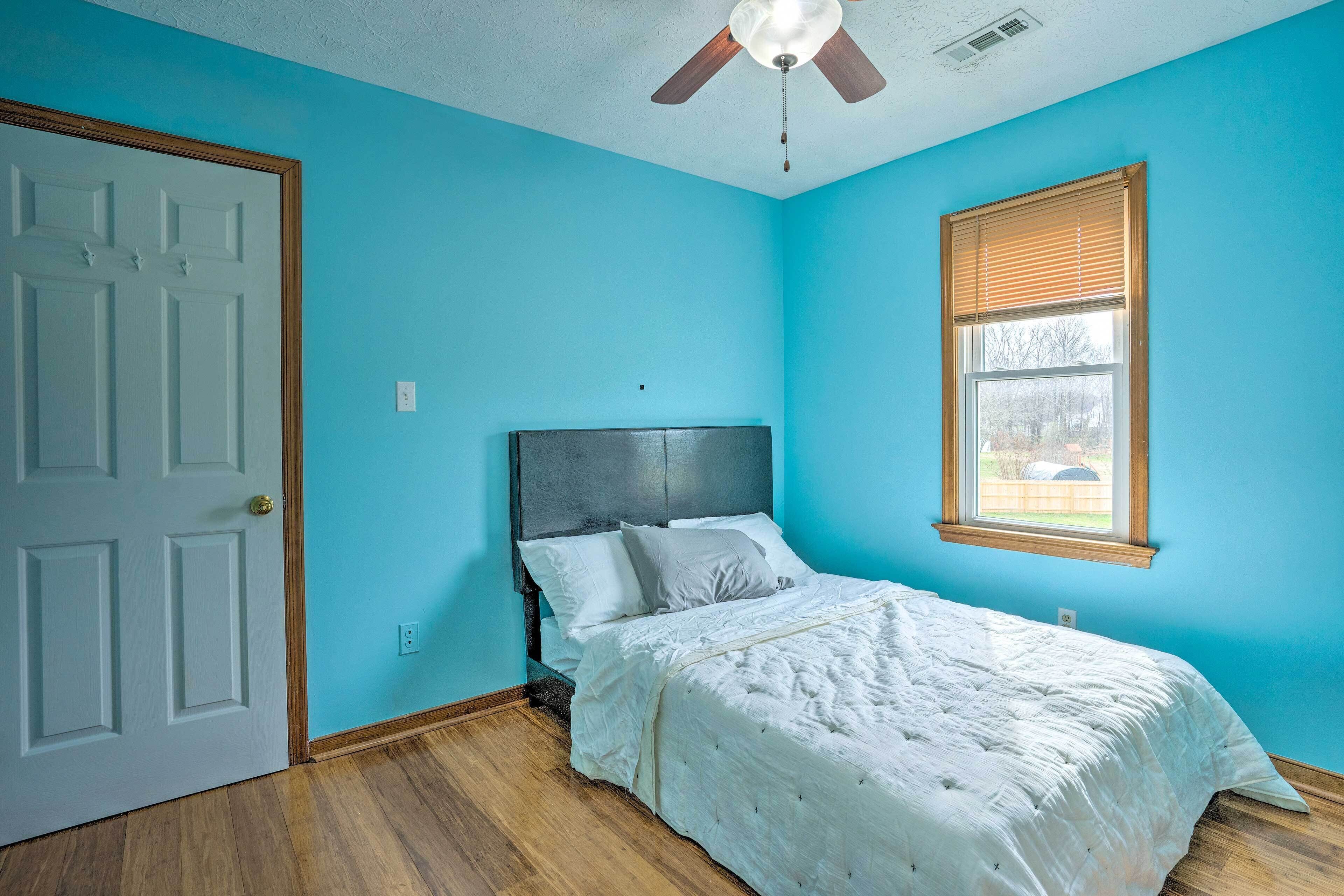 Quarto com cama, ventoinha no teto, janela com persianas e porta.