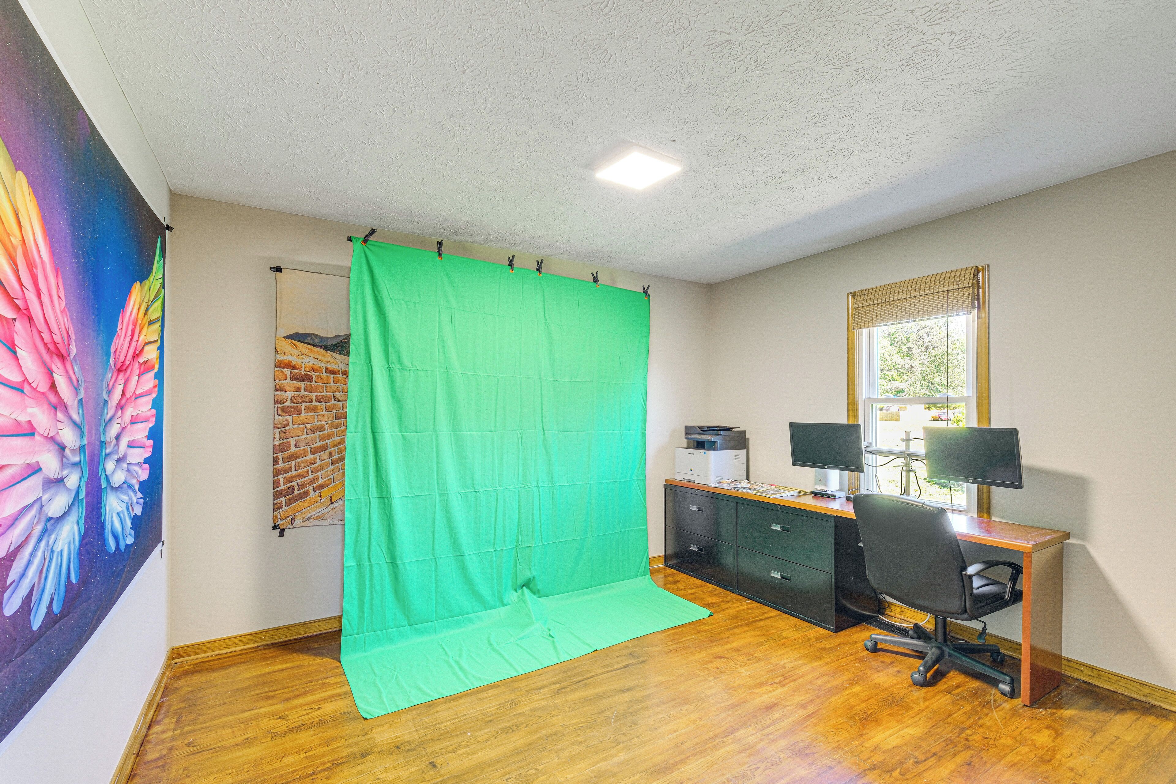 Una habitación con una pantalla verde, un escritorio con dos monitores, una silla y una ventana con persianas.