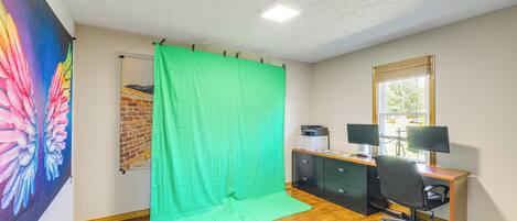 Una habitación con una pantalla verde, un escritorio con dos monitores, una silla y una ventana con persianas.