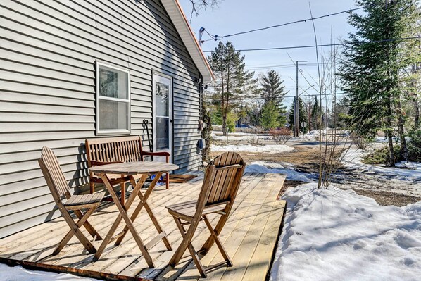 A wooden deck with a table and chairs, a window, and a view of a snowy landscape with trees.