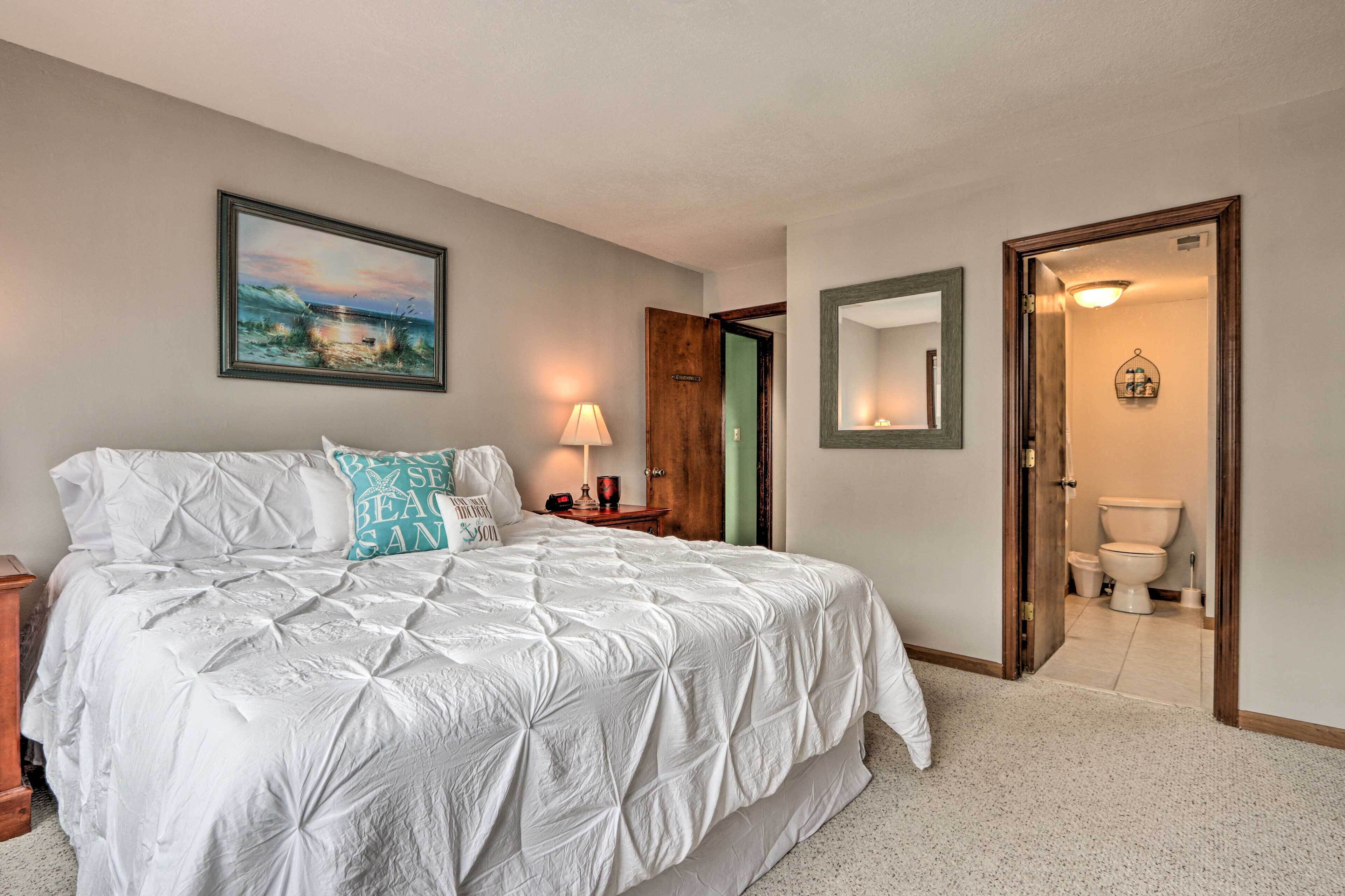 A bedroom with a large bed, a nightstand, a lamp, a painting on the wall, and a door leading to a bathroom.