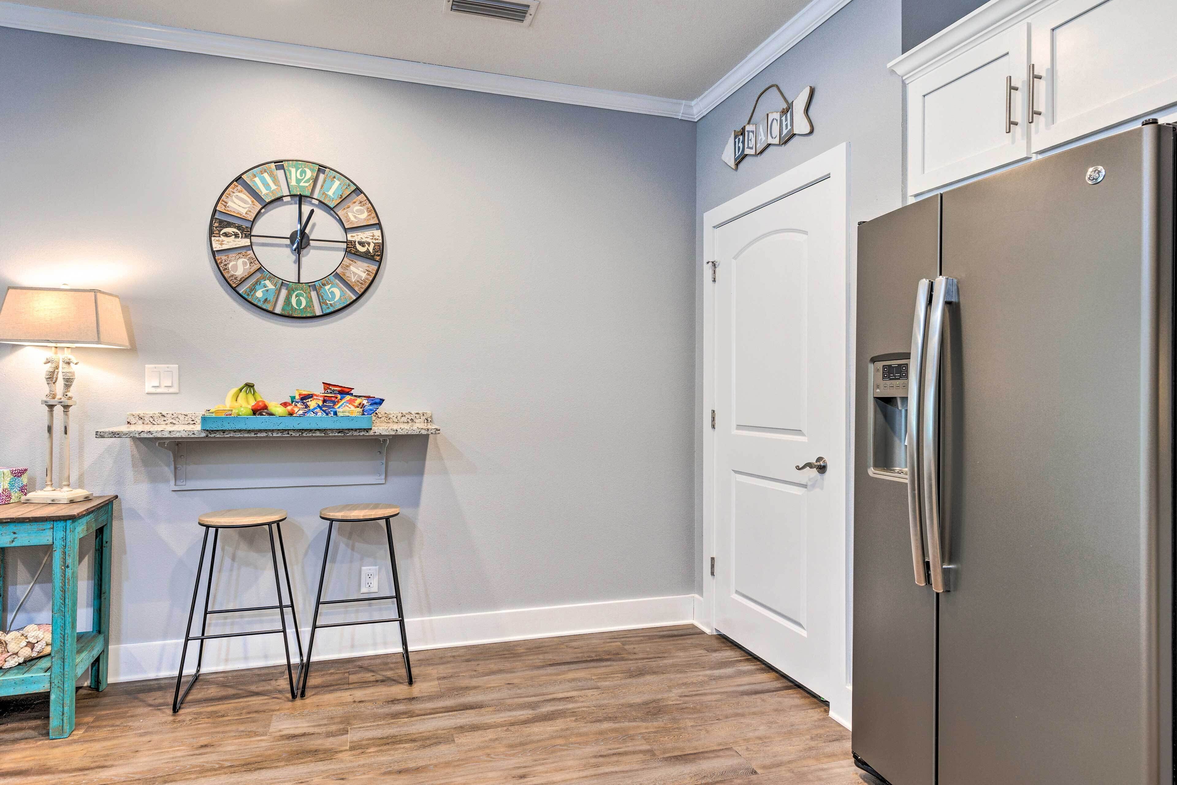 A kitchen with a stainless steel refrigerator, a clock on the wall, and two stools.
