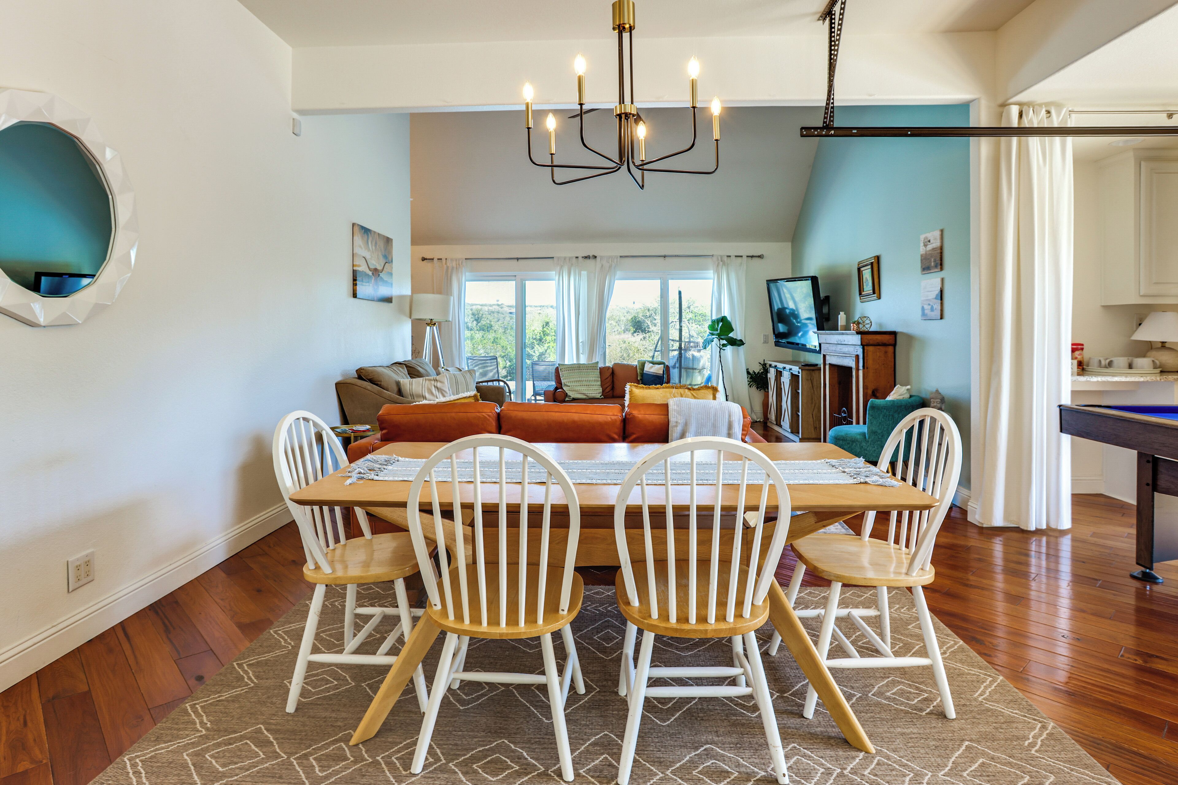 A dining area with a wooden table and white chairs, a chandelier, and a view of the outdoors through large windows.