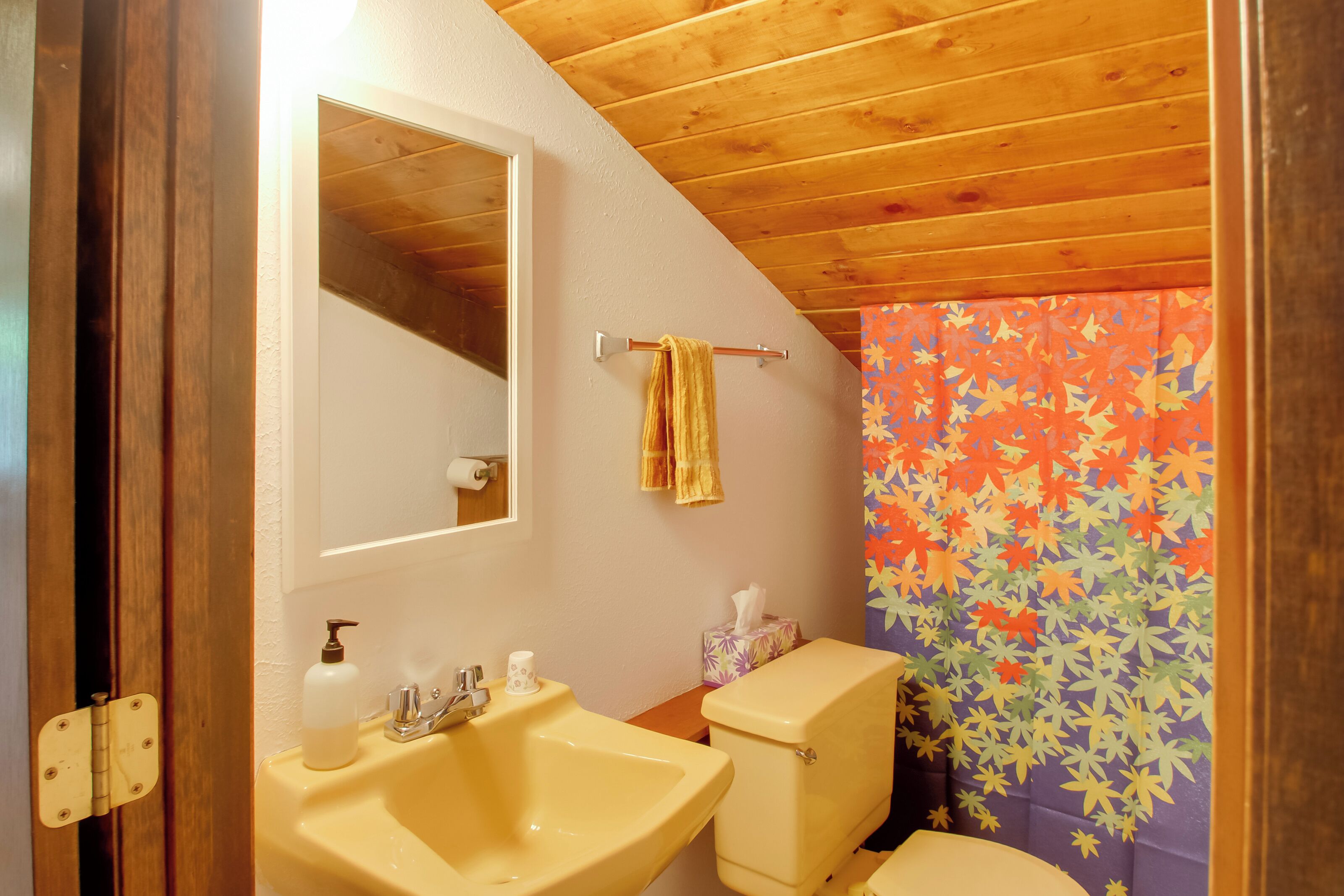 Un baño con techo de madera, un lavamanos amarillo, un inodoro y una cortina de ducha colorida.