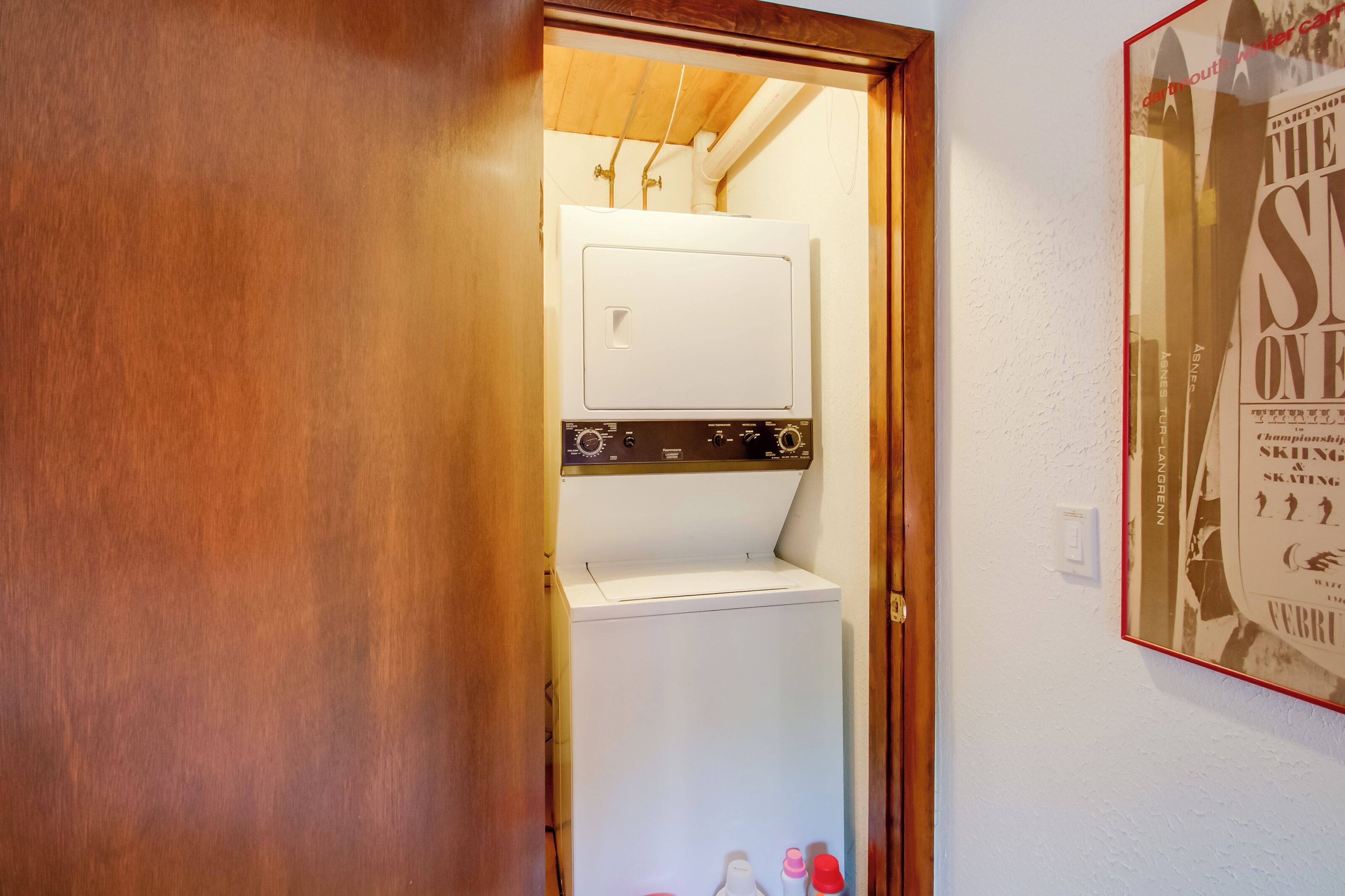 Une buanderie équipée d’un lave-linge et d’un sèche-linge, avec une porte en bois et une affiche accrochée au mur.