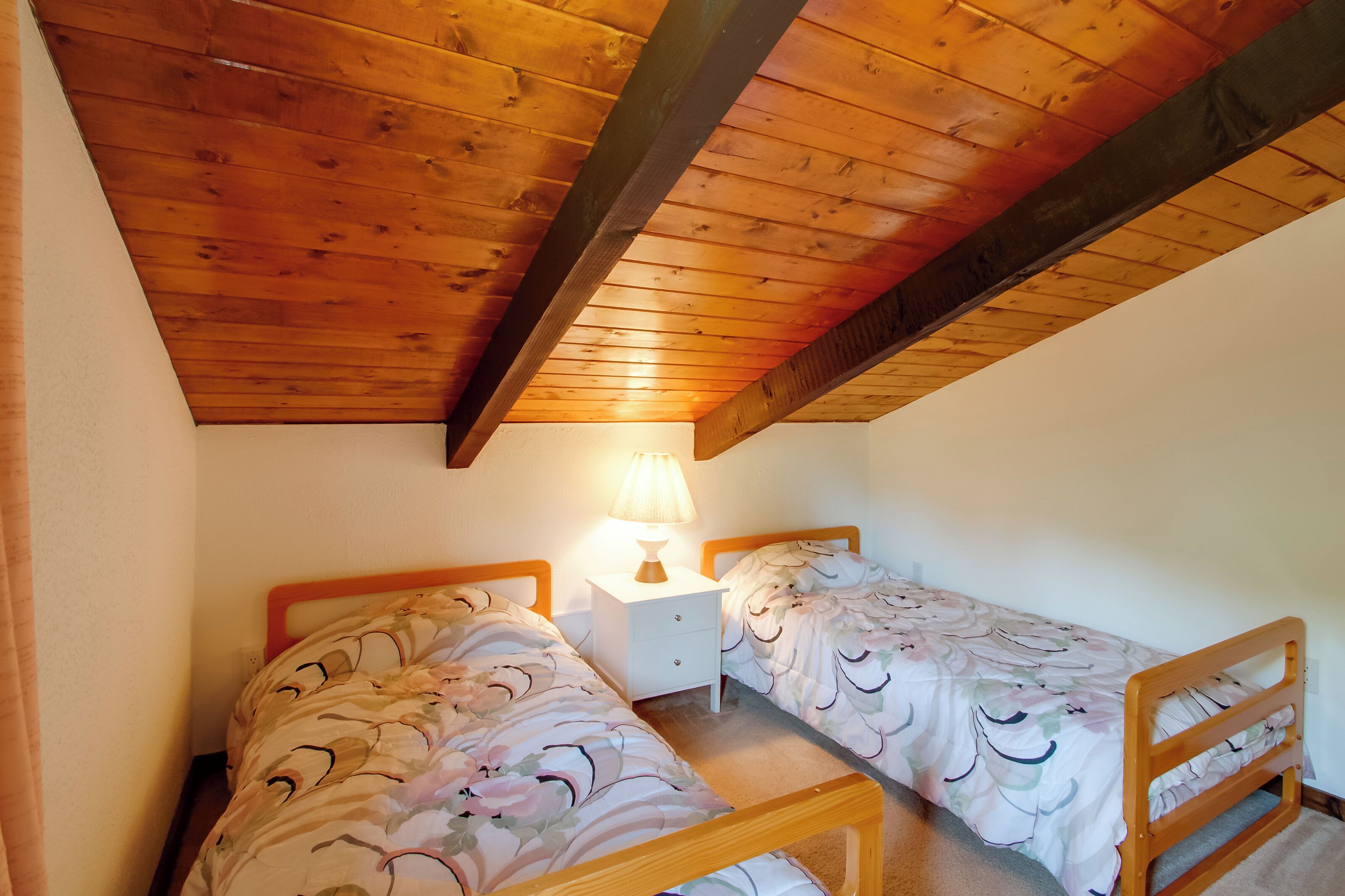 Een slaapkamer met twee eenpersoonsbedden, houten hoofdborden, een nachtkastje met een lamp en een schuin houten plafond.