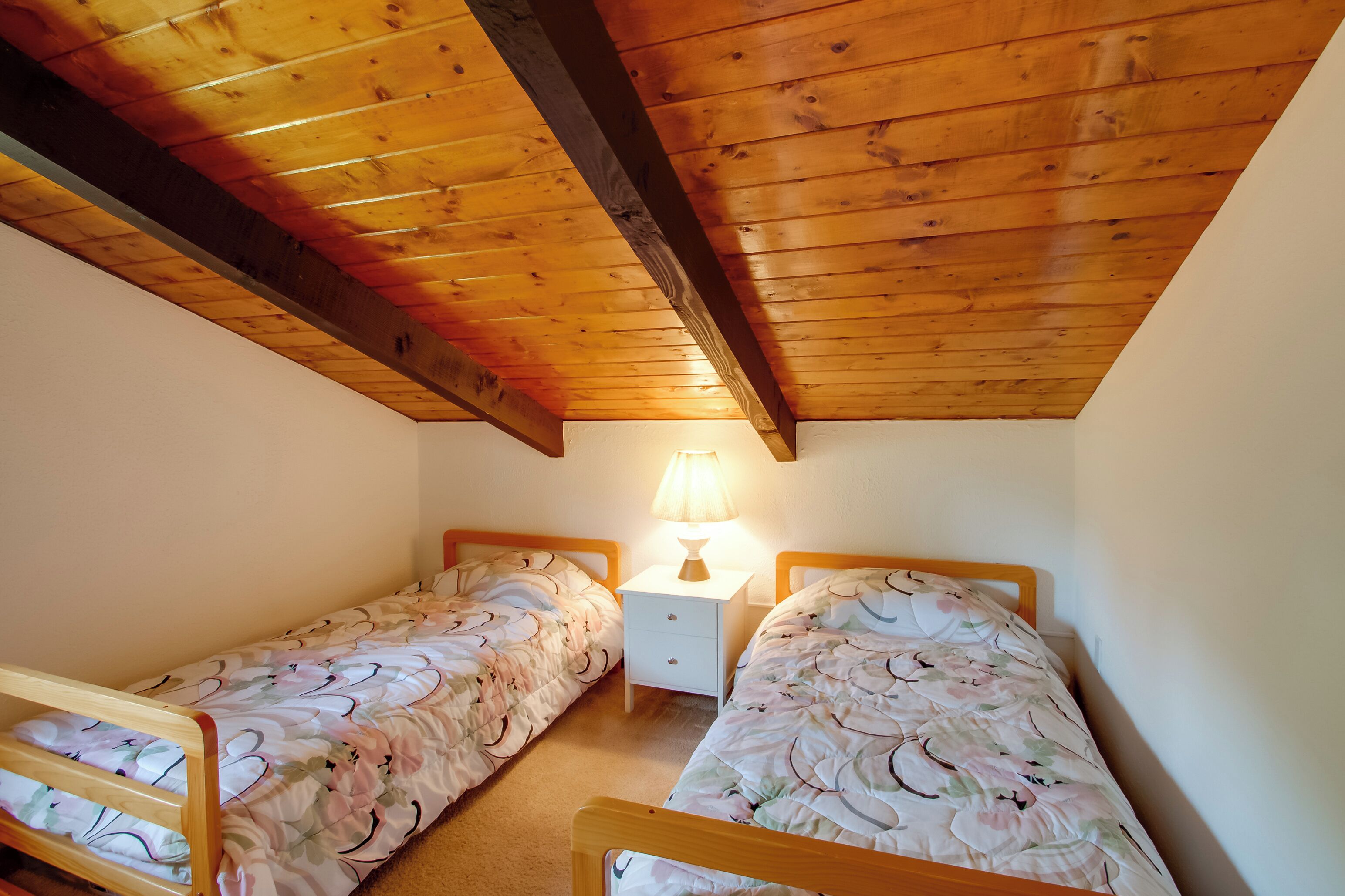 A small, cozy room with two single beds, a nightstand with a lamp, and wooden beams on the ceiling.